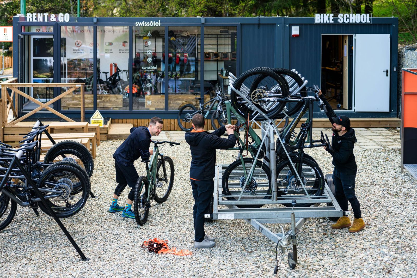 Three people working on a bike at a bike school with a building labeled 'Bike School' in the background.